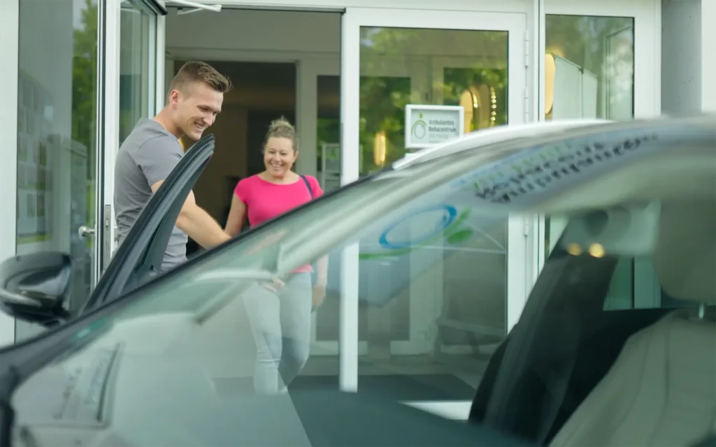 BG-Patientenfahrten 1 Vor einer Rehaeinrichtung öffnet ein Mann die Autotür, während eine Frau mit Tasche auf das Fahrzeug zugeht.